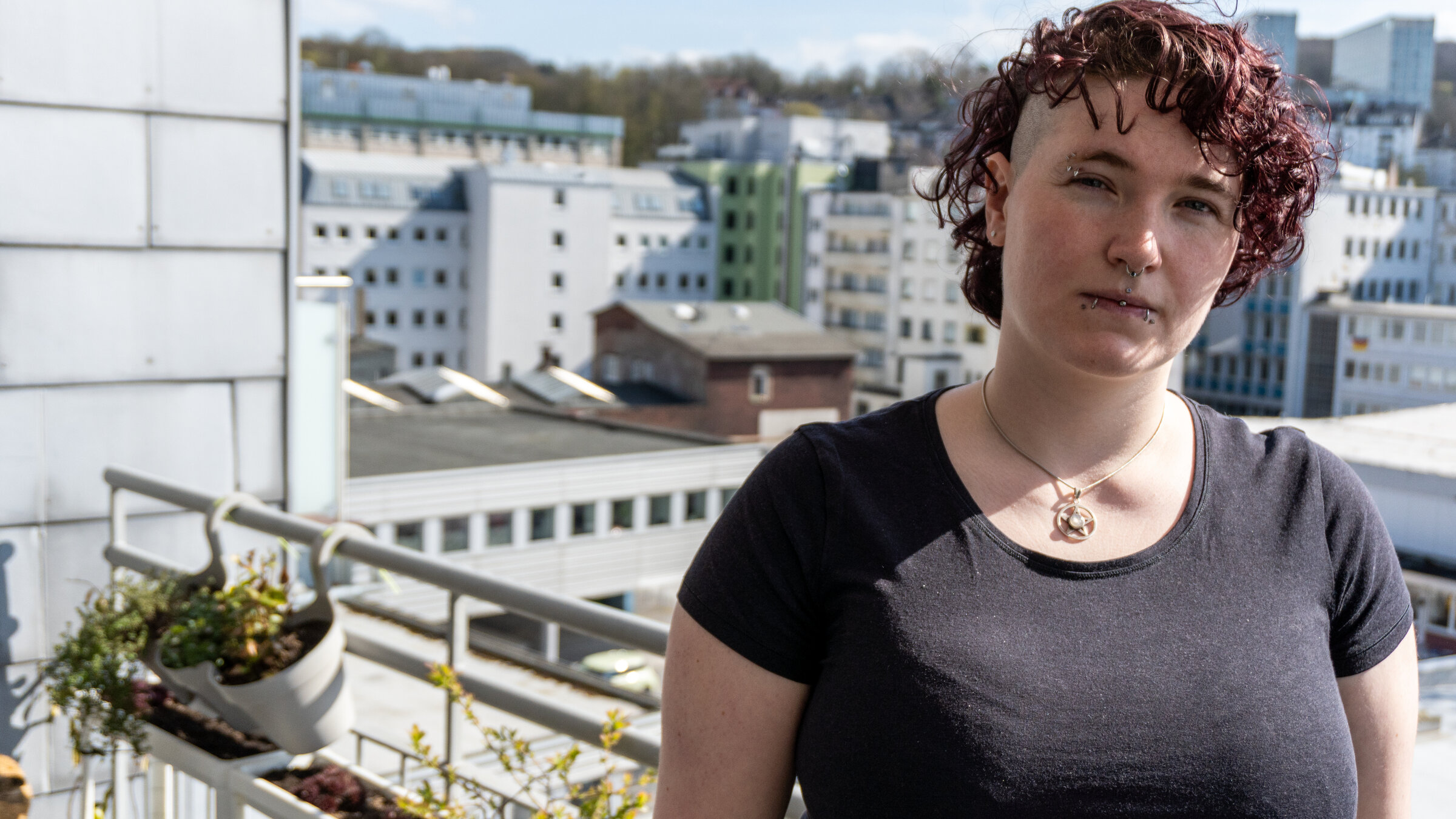 "Zwischen Not und Neustart": Genoveva (30) trägt lockiges, dunklem Haar, drei Gesichtspiercings, ein schwarzes T-Shirt. Sie steht bei sonnigem Wetter auf ihrem Balkon und blickt ruhig in die Kamera. Im Hintergrund ist die städtische Kulisse Wuppertals mit mehrstöckigen Gebäuden und begrünten Hügeln zu sehen. Auf dem Balkongeländer hängen Blumentöpfe mit Pflanzen.