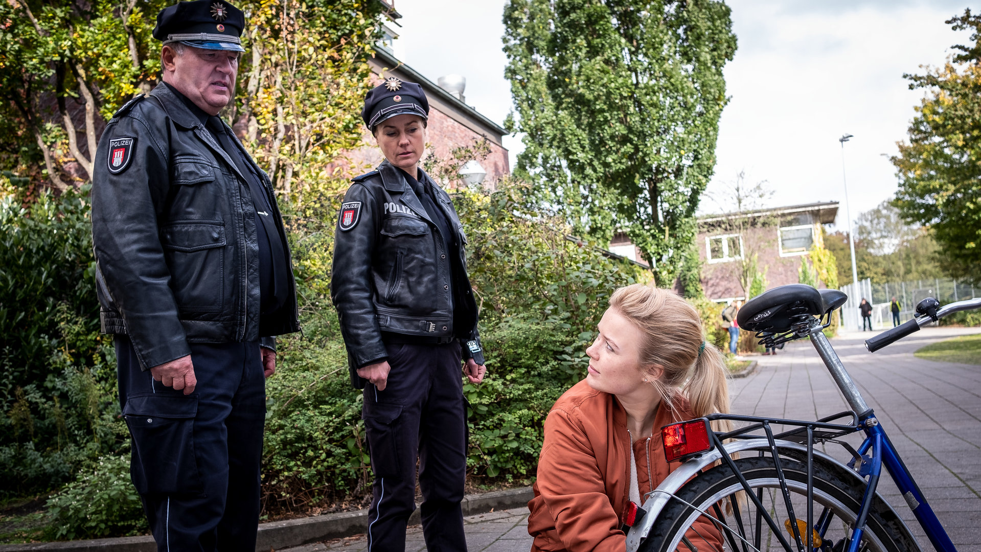 "Notruf Hafenkante - Abistreich": Der Polizist Hans Moor (Bruno F. Apitz) steht neben seiner Kollegin Franzi Jung (Rhea Harder-Vennewald) auf einem Gehweg. Beide sehen Pauline (Anna-Isabel Hinz) an, die vor ihnen neben ihrem Fahrrad kniet, das sie gerade repariert. Sie hat ihren Kopf zur Seite gerichtet und sieht zu Hans.