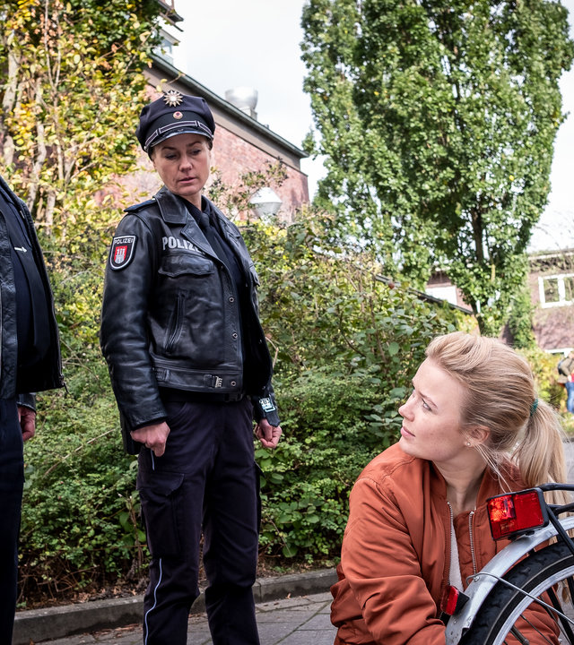 "Notruf Hafenkante - Abistreich": Der Polizist Hans Moor (Bruno F. Apitz) steht neben seiner Kollegin Franzi Jung (Rhea Harder-Vennewald) auf einem Gehweg. Beide sehen Pauline (Anna-Isabel Hinz) an, die vor ihnen neben ihrem Fahrrad kniet, das sie gerade repariert. Sie hat ihren Kopf zur Seite gerichtet und sieht zu Hans.