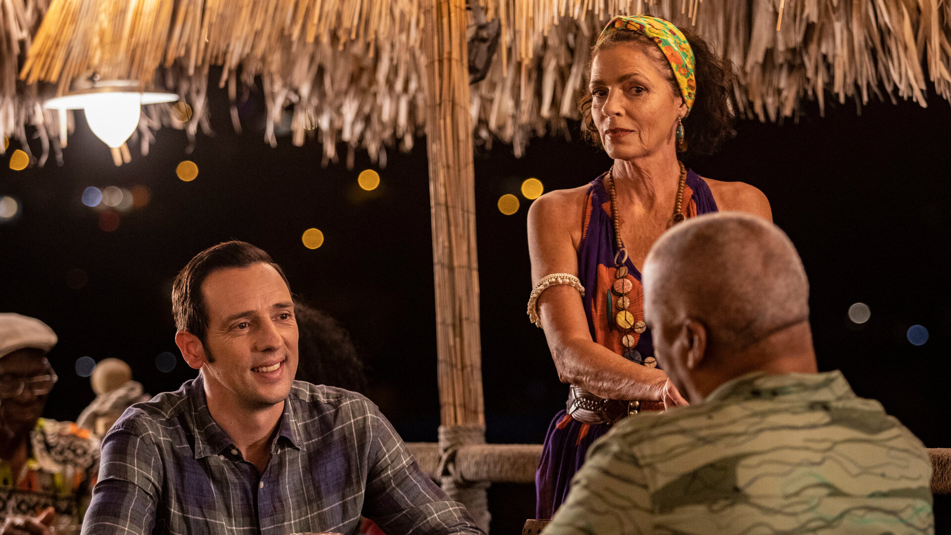 "Death in Paradise - Musik im Blut": Polizeichef Selwyn Patterson (Don Warrington) und Detective Neville Parker (Ralf Little) sitzen sich in einer Bar an einem Tisch gegenüber. Neben ihnen steht die Barfrau Catherine Bordey (Elizabeth Bourgine).