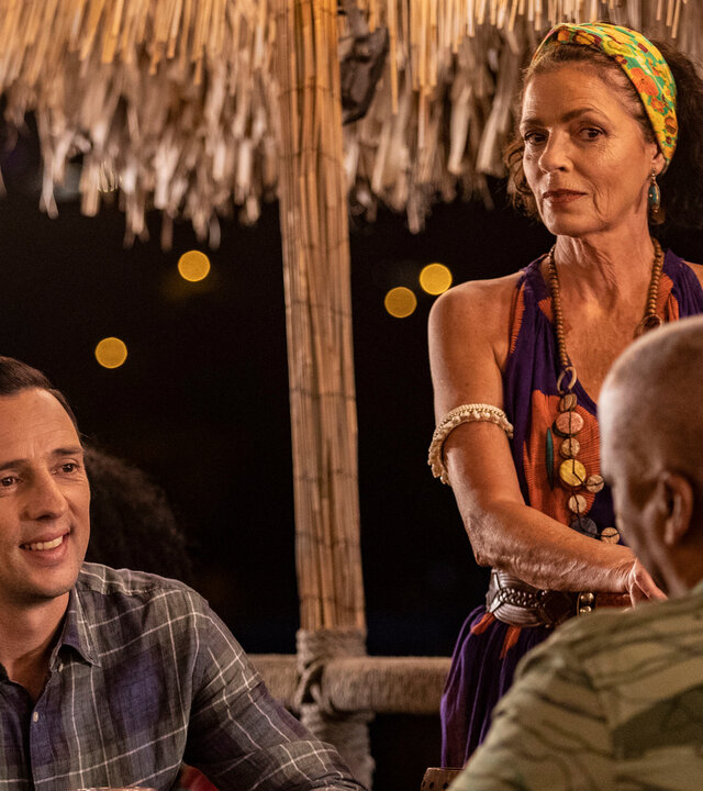 "Death in Paradise - Musik im Blut": Polizeichef Selwyn Patterson (Don Warrington) und Detective Neville Parker (Ralf Little) sitzen sich in einer Bar an einem Tisch gegenüber. Neben ihnen steht die Barfrau Catherine Bordey (Elizabeth Bourgine).