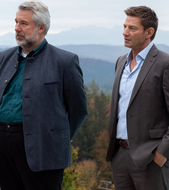 "Die Rosenheim-Cops - Was nicht passt, wird passend gemacht": Die Kommissare Stadler (Dieter Fischer) und Hansen (Igor Jeftić) stehen nebeneinander auf der Terrasse eines höher gelegenen Hauses und schauen in dieselbe Richtung. Im Hintergrund ist sind die Berge des Voralpenlandes zu sehen.