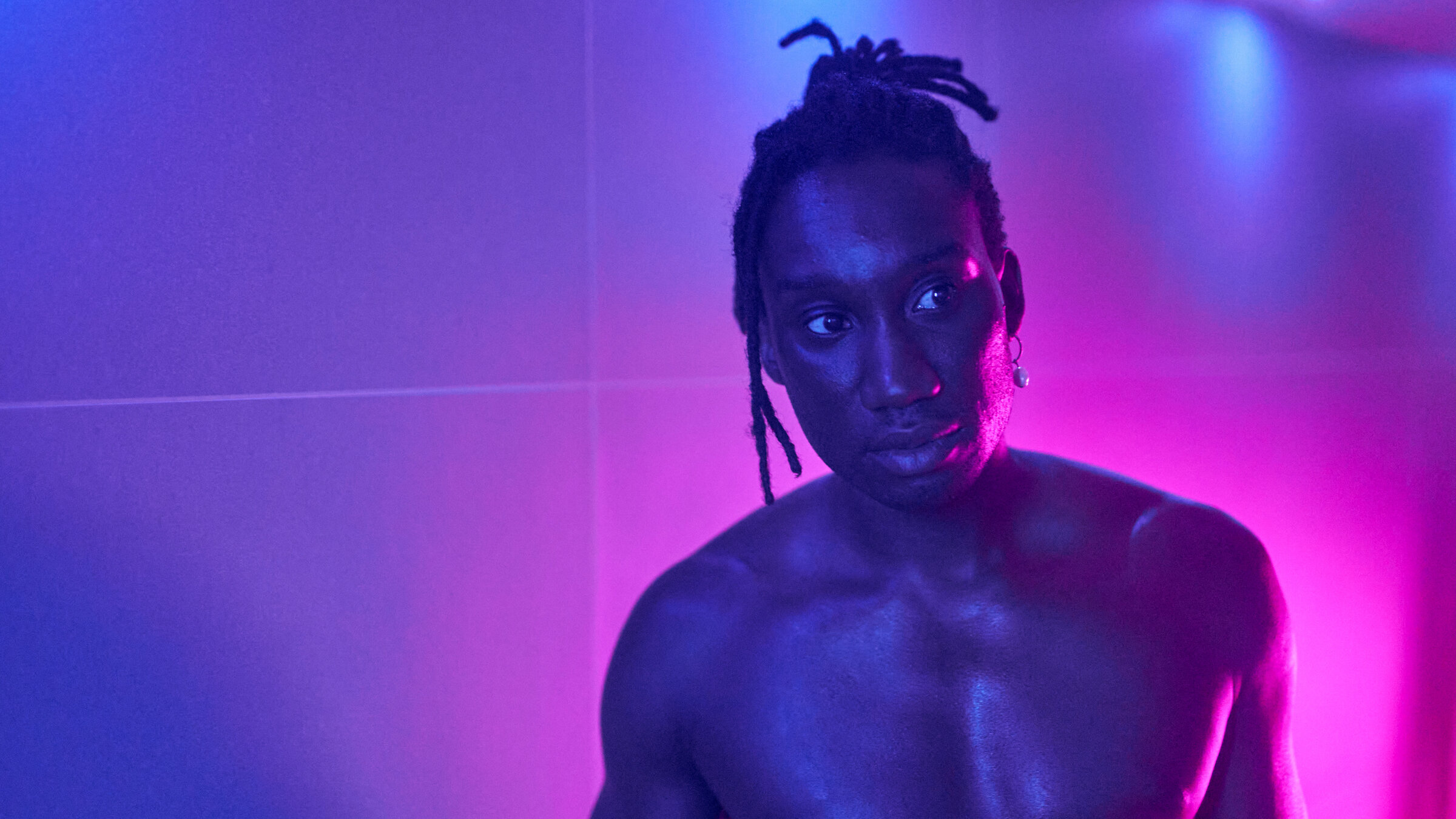 "Femme": Nahaufnahme: Jules (Nathan Stewart-Jarrett) steht oberkörperfrei vor einer gefliesten Wand in einem blau-pink ausgeleuchteten Raum. Er scheint jemanden zu beobachten.
