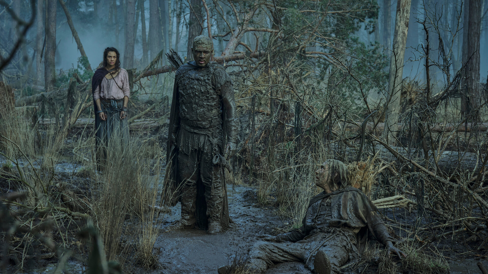 "Der Herr der Ringe: Die Ringe der Macht - Der Älteste": Isildur (Maxim Baldry) liegt in einem Moor. Arondir (Ismael Cruz Córdova) steht daneben. Beide sind von Kopf bis Fuß mit Schlamm bedeckt. Aus dem Hintergrund beobachtet die unversehrte Estrid (Nia Towle) das Geschehen. Sie trägt Handfesseln.