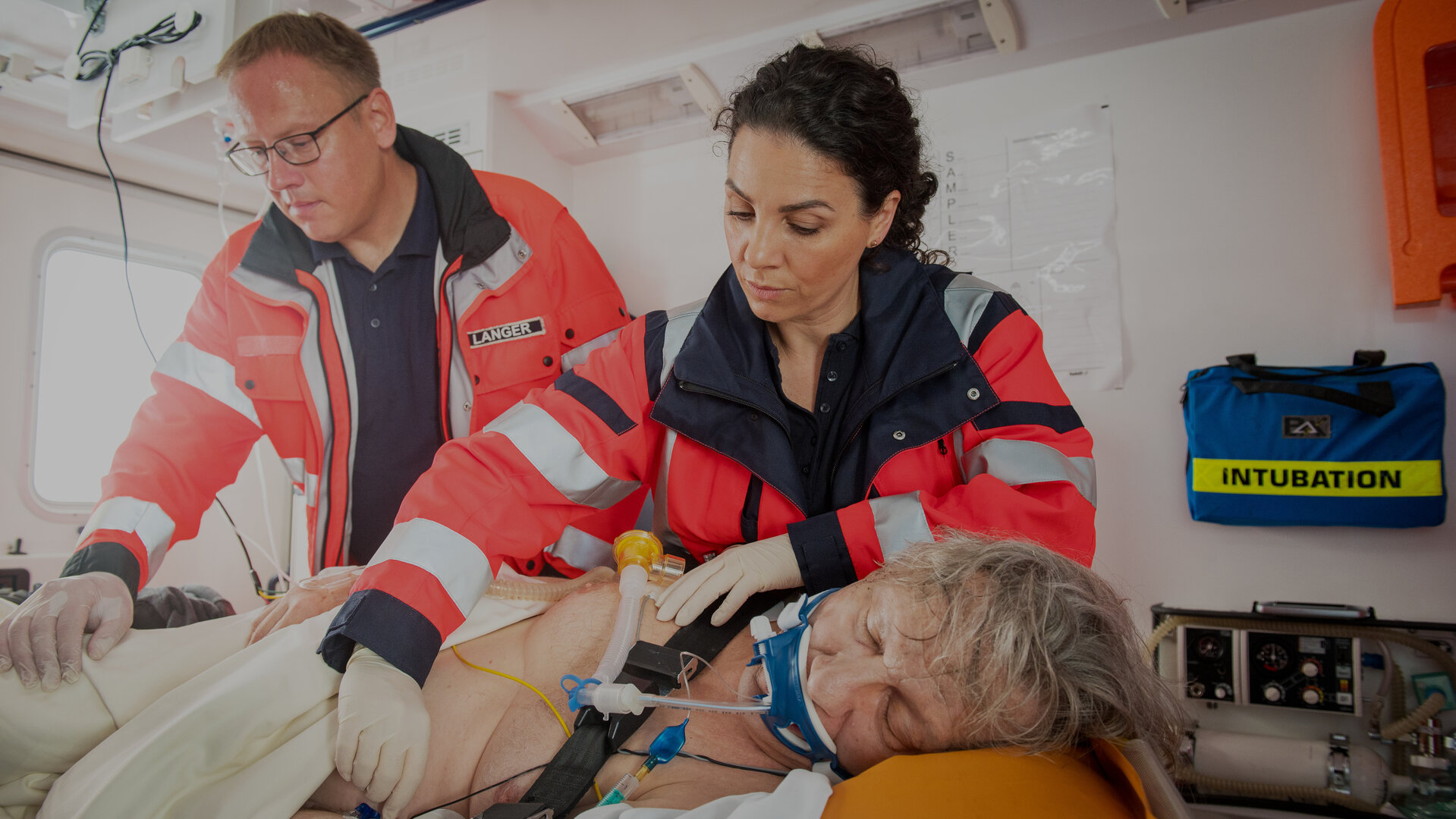 "Notruf Hafenkante - Achtung, Wolf!": Die Notärztin Dr. Sam Jordan (Hanife Sylejmani) und ein Rettungsassistenz (Komparse) stehen in einem Notarztwagen neben einer Patiententrage auf der Haller (Hannes Hellmann) liegt. Er ist auf die linke Seite gedreht und intubiert. Beide sind dabei Haller weiter zu versorgen.