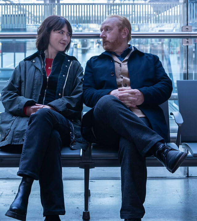 "The Chelsea Detective - Im blauen Zimmer": DS Layla Walsh (Vanessa Emme) und DI Max Arnold (Adrian Scarborough) sitzen nebeneinander auf einer Bank im Londoner Bahnhof St Pancras.