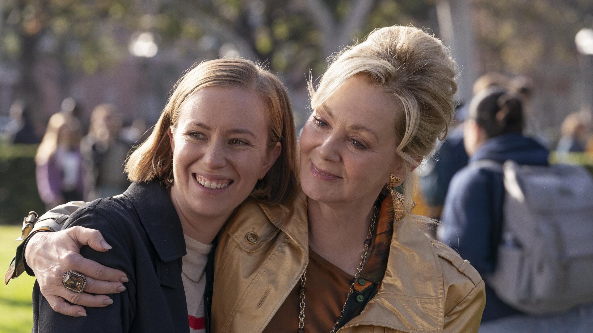 "Hacks - Niemals für einen Witz entschuldigen": Deborah (Jean Smart) lächelt und umarmt Ava Hannah Einbinder), die ebenso gelöst und glücklich lacht. Im Hintergrund befinden sich, unscharf zu erkennen, weitere Personen (Komparsen) unter Bäumen.