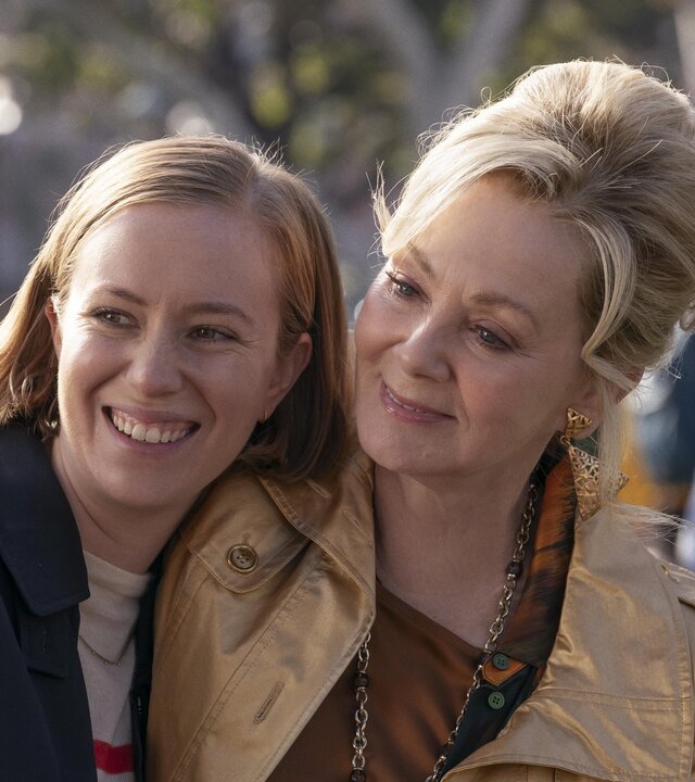 "Hacks - Niemals für einen Witz entschuldigen": Deborah (Jean Smart) lächelt und umarmt Ava Hannah Einbinder), die ebenso gelöst und glücklich lacht. Im Hintergrund befinden sich, unscharf zu erkennen, weitere Personen (Komparsen) unter Bäumen.