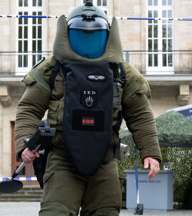 "SOKO Potsdam - Bombenstimmung": Zwei Männer (Komparsen) des SEK rücken zur Bombenentschärfung an. Ein Mann trägt eine schwere Uniform und einen Sicherheitshelm. Eine Polizistin (Komparsin) hält ihnen ein Absperrband zum Durchlaufen hoch.