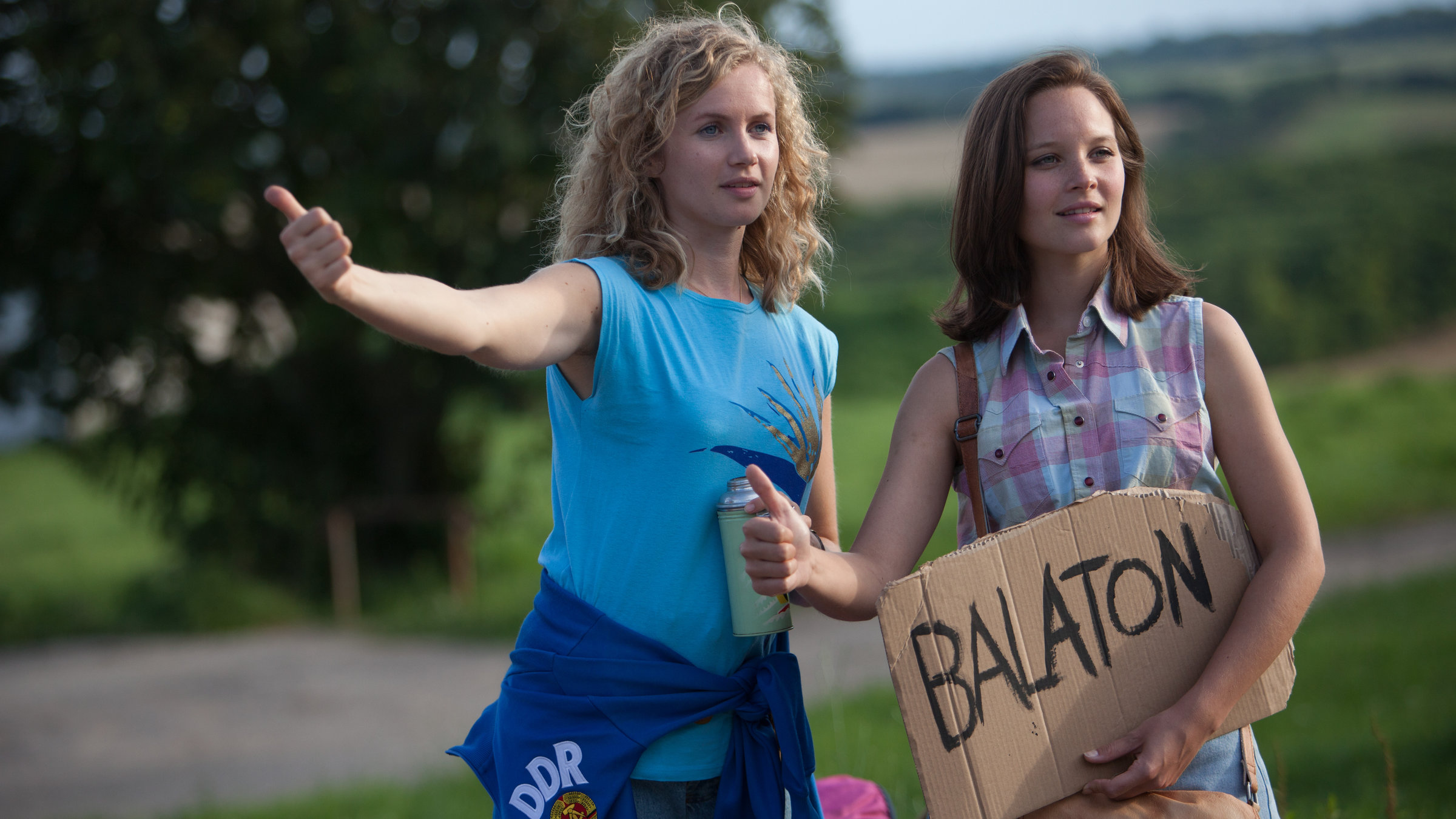 "Honigfrauen - 1. Urlaub im Paradies": Catrin Streesemann (Cornelia Gröschel) und Maja Streesemann (Sonja Gerhardt), die ein Pappschild mit der Aufschrift "Balaton" in der Hand hält, heben ihren Daumen, um per Anhalter mitgenommen zu werden.
