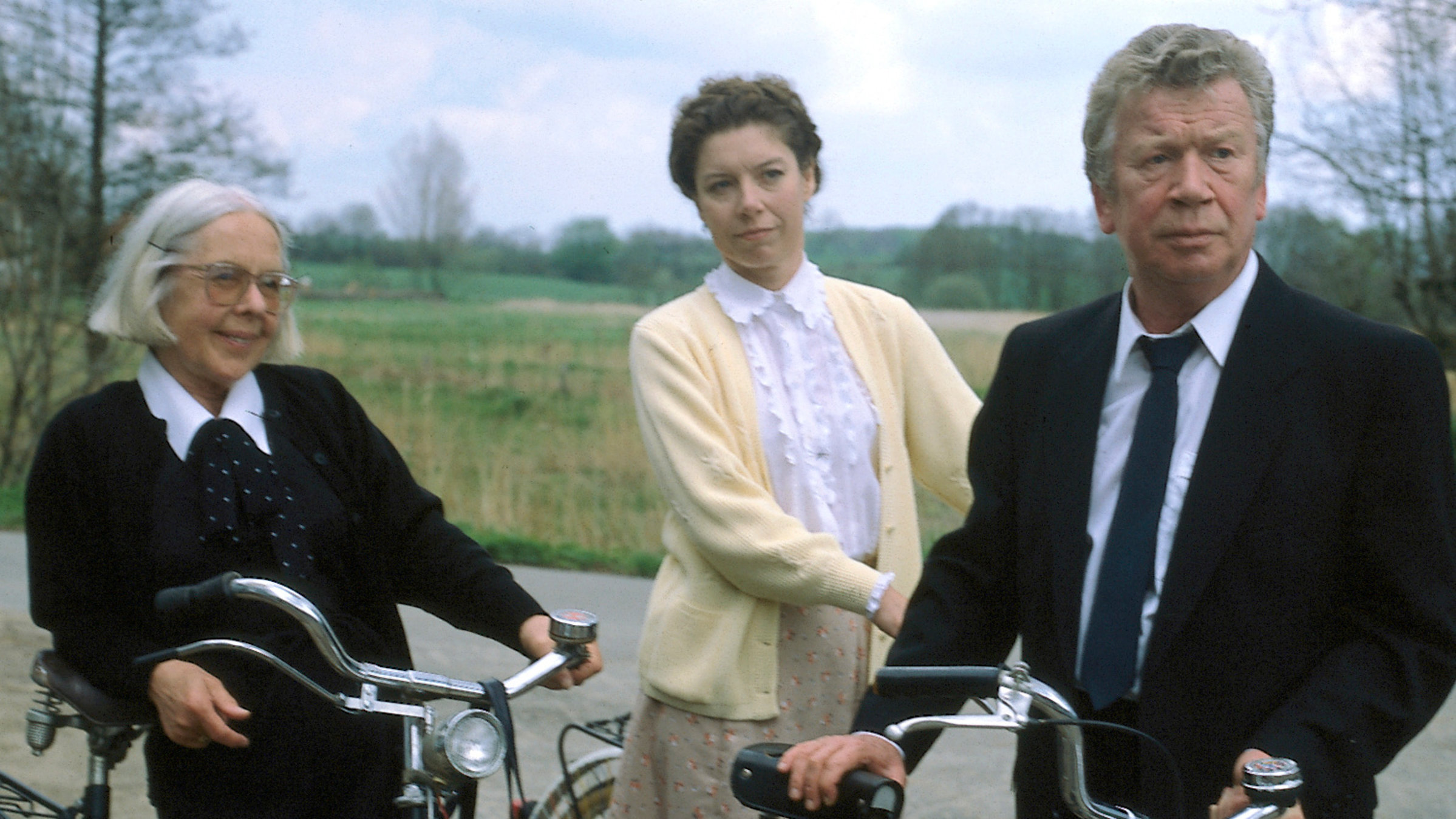 "Der Landarzt - Glücklich geschieden": Berta (Gerda Gmelin), Thea (Evelyn Hamann) und Pfarrer Eckholm (Heinz Reincke) stehen mit den Fahrrädern auf einem Landweg.