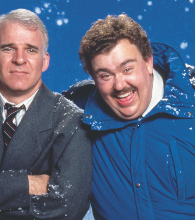 "Ein Ticket für zwei": Neal (Steve Martin, l.) und Del (John Candy, r.) sitzen nebeneinander. Del hat den rechten Arm um Neals Schulter gelegt und lacht. Neal schaut gequält. Im Hintergrund rieseln Schneeflocken.