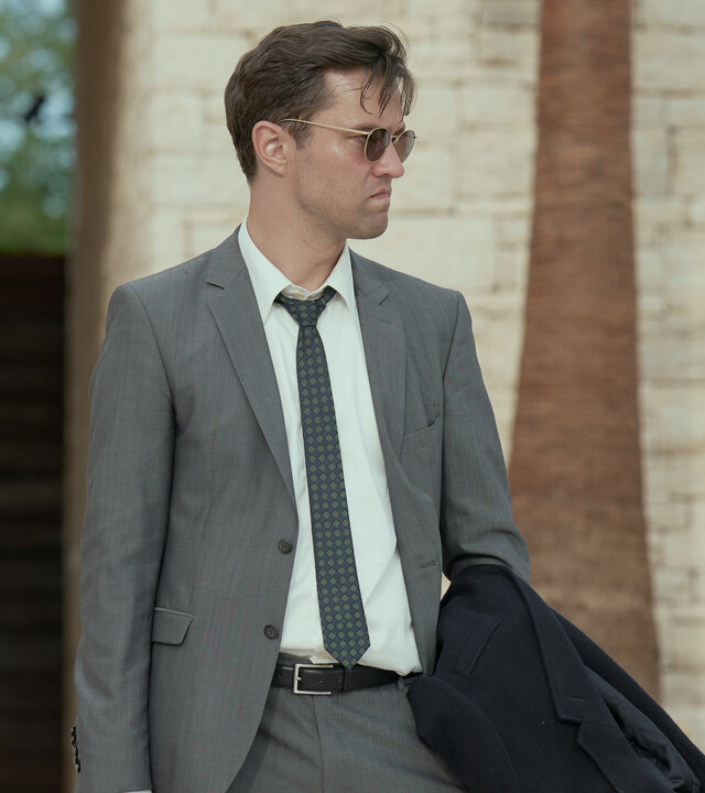 "Die Affäre Cum-Ex - Episode 3": Im sonnigen Garten einer Villa geht Sven Lebert (Nils Strunk) in Anzug und mit Sonnenbrille auf die Kamera zu. Fred Allen (Andy Murray) trägt ein offenes Hemd und zeigt seinen muskulösem Oberkörper. Er folgt Fred, während eine Frau (Komparsin) sich im Hintergrund der Sonne entgegen streckt.