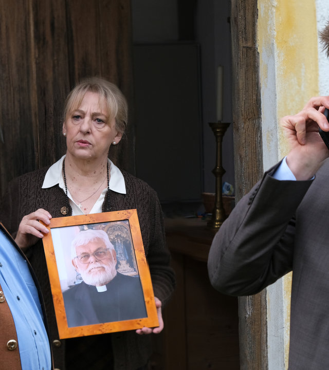 "Die Rosenheim-Cops - Kein toter Pfarrer": Die Kommissare Stadler (Dieter Fischer) und Hansen (Igor Jeftić) stehen vor der Tür der Pfarrei. Im Türrahmen steht die Haushälterin Gerda Schlobinger (Johanna Bittenbinder), die ein gerahmtes Portrait eines Pfarrers in den Händen hält, während sie Stadler ansieht. Dieser spricht mit Hansen, der gerade telefoniert.