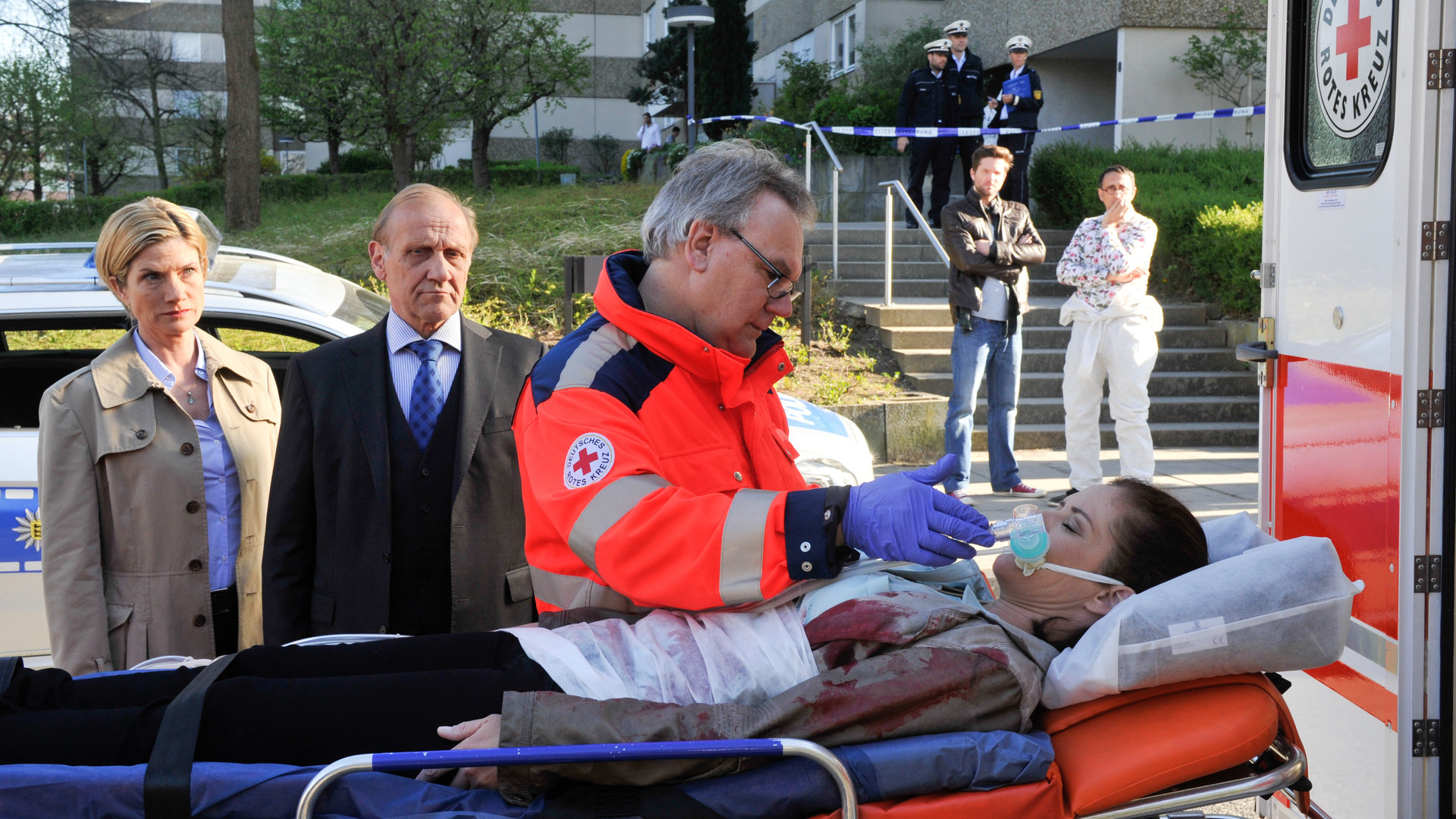 "SOKO Stuttgart - Das Gesetz der Straße": Selma Kirsch (Yve Burbach) ist mit einem Messerstich in die Herzgegend schwer verletzt worden. Sie liegt auf der Trage, neben ihr steht der Notarzt (Komparse). Martina Seiffert (Astrid M. Fünderich) und Michael Kaiser (Karl Kranzkowski) stehen tief bestürzt daneben. Etwas weiter weg stehen genauso entsetzt Jo Stoll (Peter Ketnath) und Jan Arnaud (Mike Zaka Sommerfeldt).