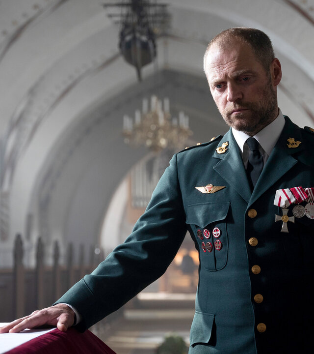 "Oxen - Für Ehre und Vaterland": Niels Oxen (Jacob Lohmann) steht in einer Kirche und legt die rechte Hand auf einen Sarg. Er trägt seine Uniform mit zahlreichen Orden.