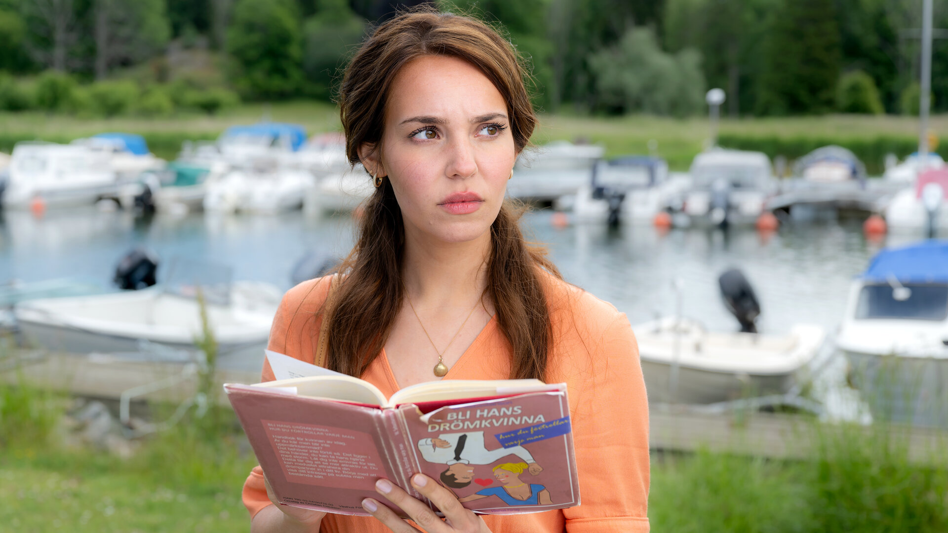 "Inga Lindström: Ein Traummann zum Üben": Finja (Ina Hout) steht am Hafen und blickt vom aufgeschlagenen Beziehungsratgeber auf. Im Hintergrund liegen vertäute Boote im Hafen.