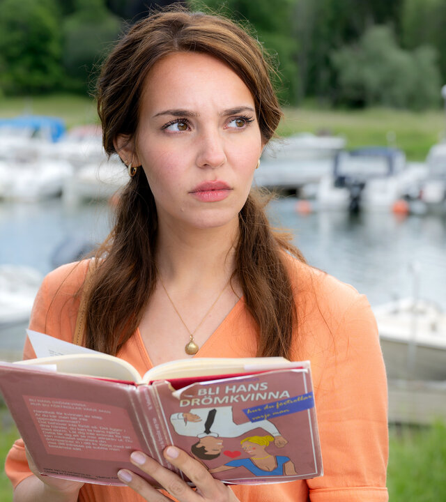 "Inga Lindström: Ein Traummann zum Üben": Finja (Ina Hout) steht am Hafen und blickt vom aufgeschlagenen Beziehungsratgeber auf. Im Hintergrund liegen vertäute Boote im Hafen.