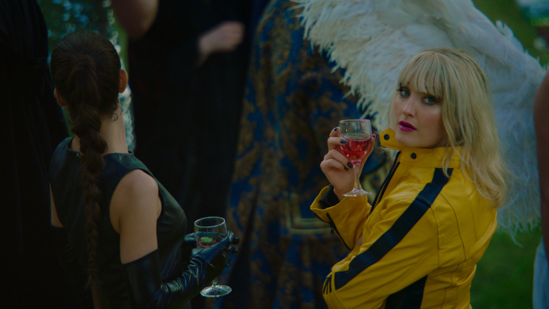 "Exit - Maskenball": Celine (Ine Wilmann) und Hermine (Agnes Kittelsen) stehen kostümiert inmitten einer Menschenmenge. Während Hermine die anderen Personen ansieht, blickt Celine mit einem Drink in ihrer Hand über ihre Schulter in Richtung Kamera.