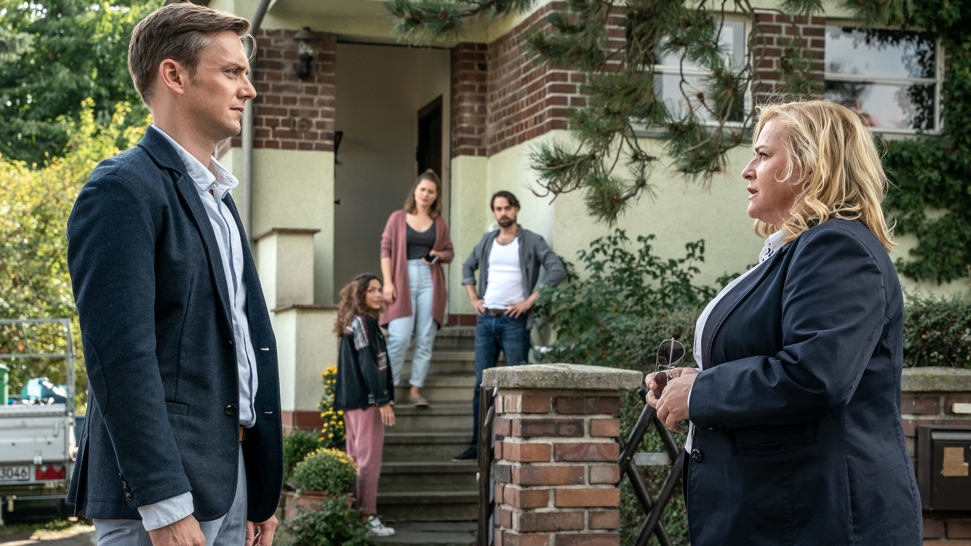 "SOKO Leipzig - Betrogen": Tobias Koch (Jan Dose) steht im Vordergrund mit Dagmar Schnee (Petra Kleinert) in einer Hauseinfahrt. Kim Nowak (Amy Mußul), Iris Michalski (Laura Lippmann) und Jonas Michalski (Fabian Oehl) stehen im Hintergrund auf der Treppe eines Hauses und beobachten das Geschehen im Vordergrund.