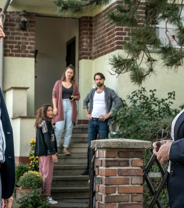 "SOKO Leipzig - Betrogen": Tobias Koch (Jan Dose) steht im Vordergrund mit Dagmar Schnee (Petra Kleinert) in einer Hauseinfahrt. Kim Nowak (Amy Mußul), Iris Michalski (Laura Lippmann) und Jonas Michalski (Fabian Oehl) stehen im Hintergrund auf der Treppe eines Hauses und beobachten das Geschehen im Vordergrund.
