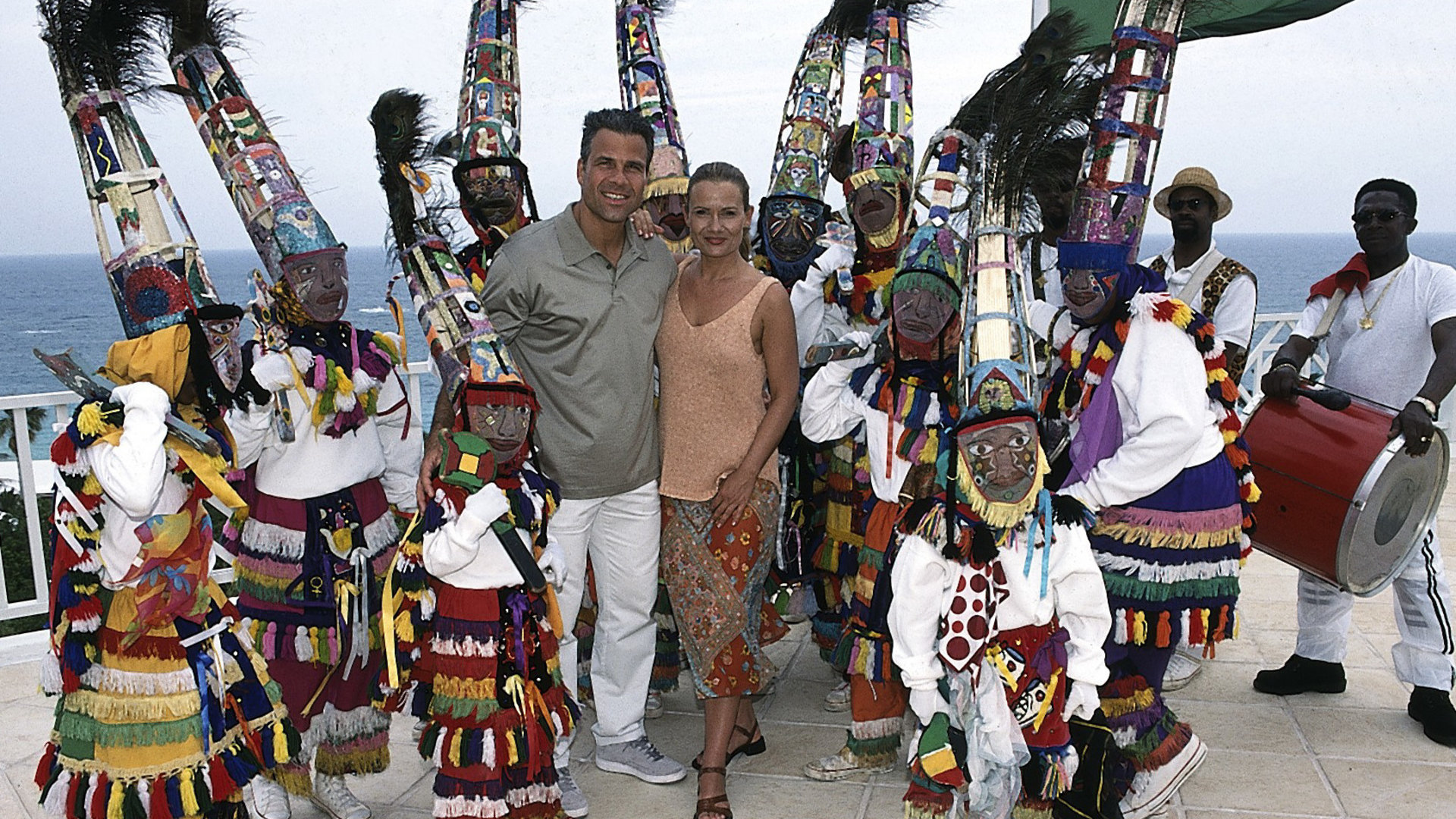 "Das Traumschiff: Bermuda": Daniel (Karsten Speck) und Viola (Jenny Jürgens) stehen Arm in Arm nebeneinander auf einer Terrasse und lächeln in die Kamera. Um sie herum stehen Musiker und eine Tanzgruppe in bunten Gewändern und Hüten. Sie tragen zusätzlich noch Masken. Im Hintergrund sieht man das Meer.
