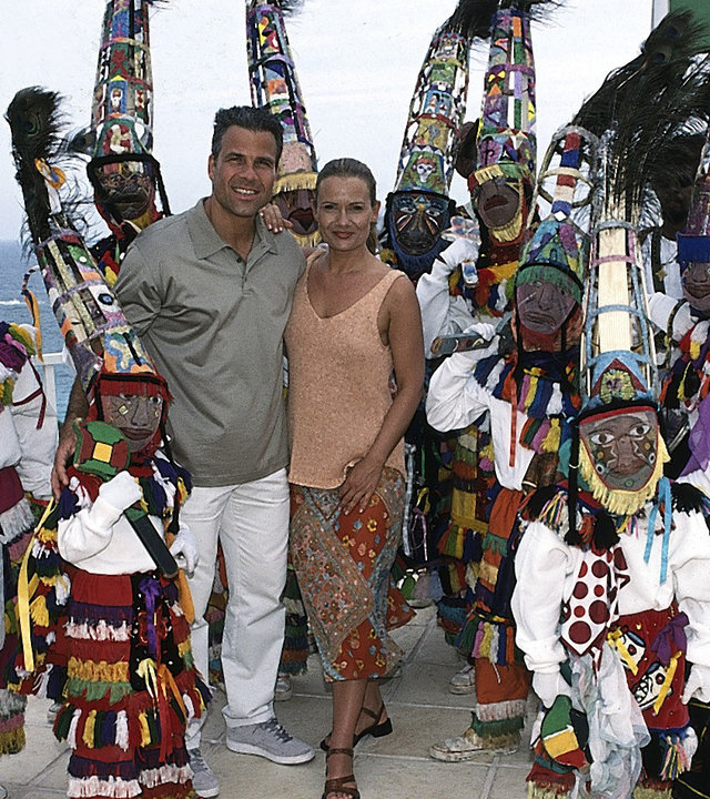 "Das Traumschiff: Bermuda": Daniel (Karsten Speck) und Viola (Jenny Jürgens) stehen Arm in Arm nebeneinander auf einer Terrasse und lächeln in die Kamera. Um sie herum stehen Musiker und eine Tanzgruppe in bunten Gewändern und Hüten. Sie tragen zusätzlich noch Masken. Im Hintergrund sieht man das Meer.