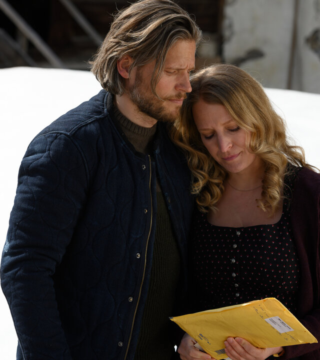 "Die Bergretter: Letzter Wille": Außenaufnahme an einer Hütte, davor, fast mannshoch, aufgehäufter Schnee. Markus (Sebastian Ströbel) hält Emilie (Stefanie von Poser) im Arm, die einen gelben Briefumschlag in der Hand hält und traurig darauf blickt.