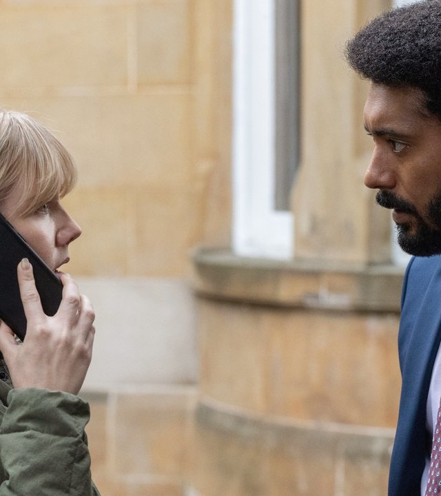 "Karen Pirie - Echo einer Mordnacht - Späte Sühne": Karen (Lauren Lyle) steht telefonierend vor dem Haus und schaut Phil (Zach Wyatt) an, der vor ihr steht.