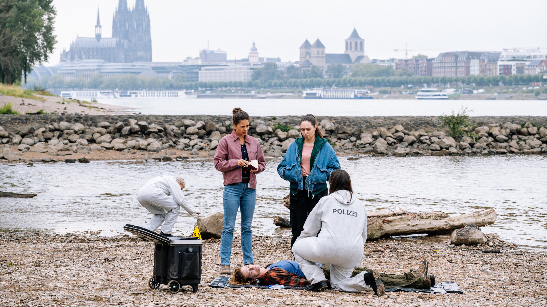 "SOKO Köln - Scherben bringen Unglück": Am Rheinufer mit Blick auf den Kölner Dom stehen Lilly Funke (Tatjana Kästel) und Amira Mahdi (Aylin Ravanyar ) neben Christine Sperling (Julia Doege), die auf dem sandigen Boden auf dem Rücken liegt. Lilly hält ein aufgeschlagenes Notizbuch in den Händen. Amira hat die Hände in den Taschen ihrer Jeansjacke vergraben. Beide blicken nachdenklich zu der Toten. Dr. Westphal (Yulia Yáñez Schmidt) kniet mit dem Rücken zur Kamera vor Christine Sperling und sieht nach oben zu den Kommissarinnen. Sie trägt einen weißen Schutzanzug mit der Aufschrift "Polizei". Im Hintergrund untersucht ein Mitarbeiter der Spurensicherung einen Stein.
