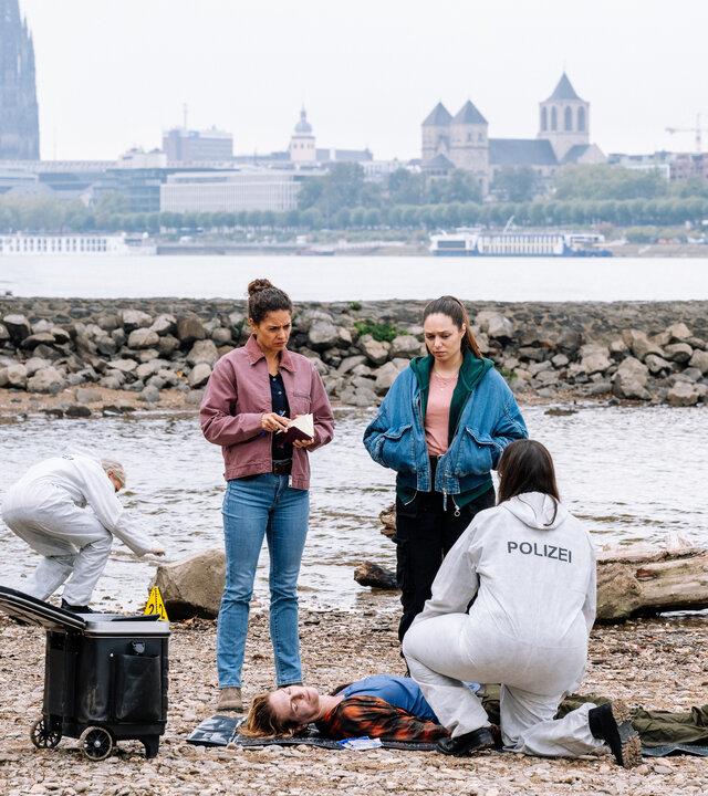 "SOKO Köln - Scherben bringen Unglück": Am Rheinufer mit Blick auf den Kölner Dom stehen Lilly Funke (Tatjana Kästel) und Amira Mahdi (Aylin Ravanyar ) neben Christine Sperling (Julia Doege), die auf dem sandigen Boden auf dem Rücken liegt. Lilly hält ein aufgeschlagenes Notizbuch in den Händen. Amira hat die Hände in den Taschen ihrer Jeansjacke vergraben. Beide blicken nachdenklich zu der Toten. Dr. Westphal (Yulia Yáñez Schmidt) kniet mit dem Rücken zur Kamera vor Christine Sperling und sieht nach oben zu den Kommissarinnen. Sie trägt einen weißen Schutzanzug mit der Aufschrift "Polizei". Im Hintergrund untersucht ein Mitarbeiter der Spurensicherung einen Stein.