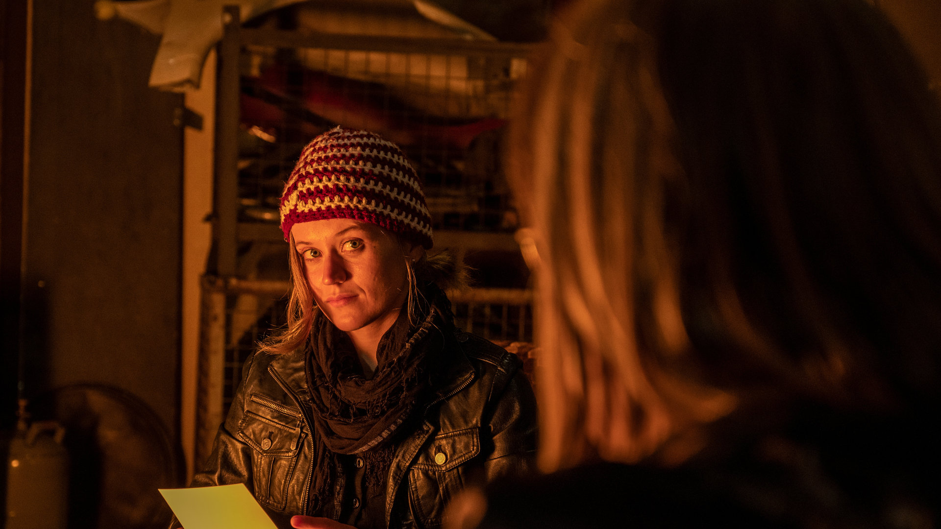 "SOKO Stuttgart – Vater gesucht": Sam Fitz (Camille Dombrowsky) sitzt mit Strickmütze und Lederjacke im Schein eines Lagerfeuers vor Schrottis Werkstatt. Auf ihren Knien hat sie eine Metallkiste und in den Händen hält sie eine Fotografie, die man jedoch nicht sehen kann. Sie schaut eindringlich zu Schrotti (Michael Gaedt), der verschwommen im Vordergrund zu sehen ist.