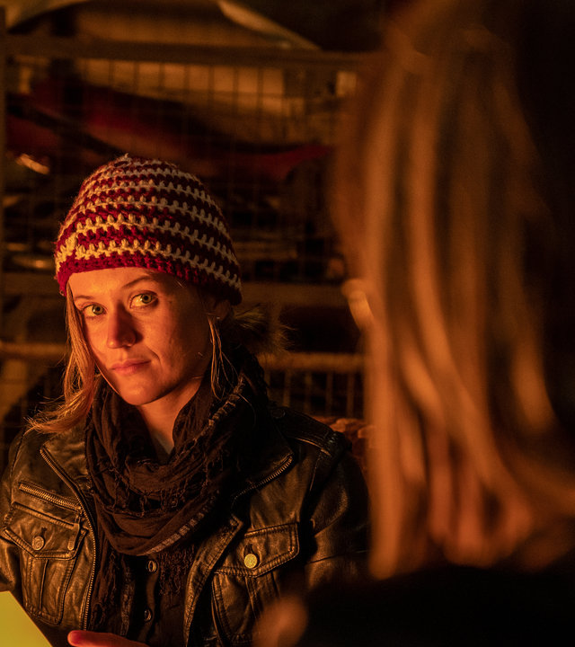 "SOKO Stuttgart – Vater gesucht": Sam Fitz (Camille Dombrowsky) sitzt mit Strickmütze und Lederjacke im Schein eines Lagerfeuers vor Schrottis Werkstatt. Auf ihren Knien hat sie eine Metallkiste und in den Händen hält sie eine Fotografie, die man jedoch nicht sehen kann. Sie schaut eindringlich zu Schrotti (Michael Gaedt), der verschwommen im Vordergrund zu sehen ist.