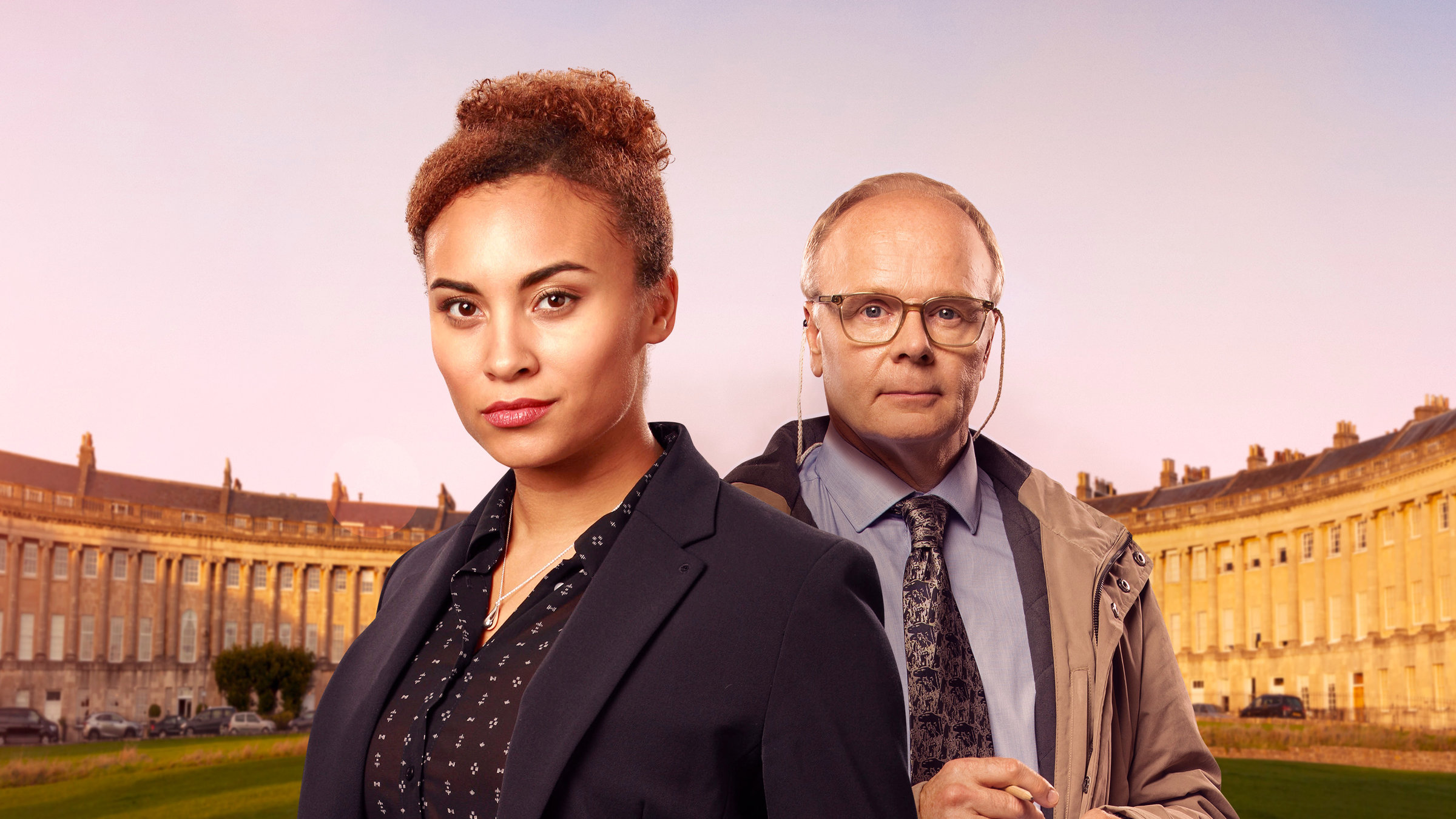 "McDonald & Dodds - Der Tote nahm den Hut": DS Dodds (Jason Watkins) und DCI Lauren McDonald (Tala Gouveia) stehen auf einem Rasen vor dem halbmondförmigen Gebäudekomplex Royal Crescent, eine Anordnung von 30 Reihenhäusern im englischen Bath.