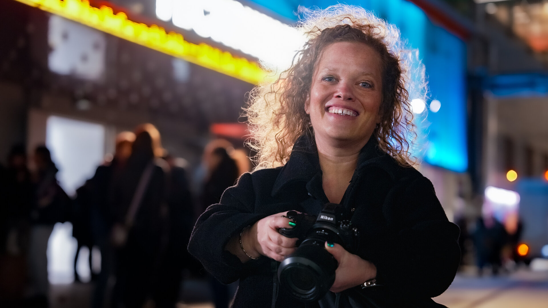 "einfach Mensch: Käthe de Koe: Kunst im Fokus": Itje Kleinert steht vor einer leuchtenden Konzerthalle und lacht in die Kamera. Dabei hält sie ihre Fotokamera mit beiden Händen fest.