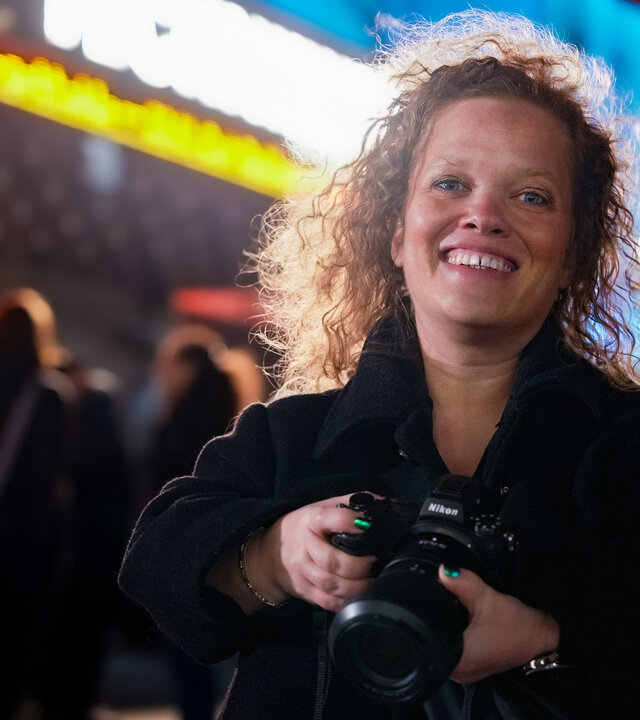 "einfach Mensch: Käthe de Koe: Kunst im Fokus": Itje Kleinert steht vor einer leuchtenden Konzerthalle und lacht in die Kamera. Dabei hält sie ihre Fotokamera mit beiden Händen fest.