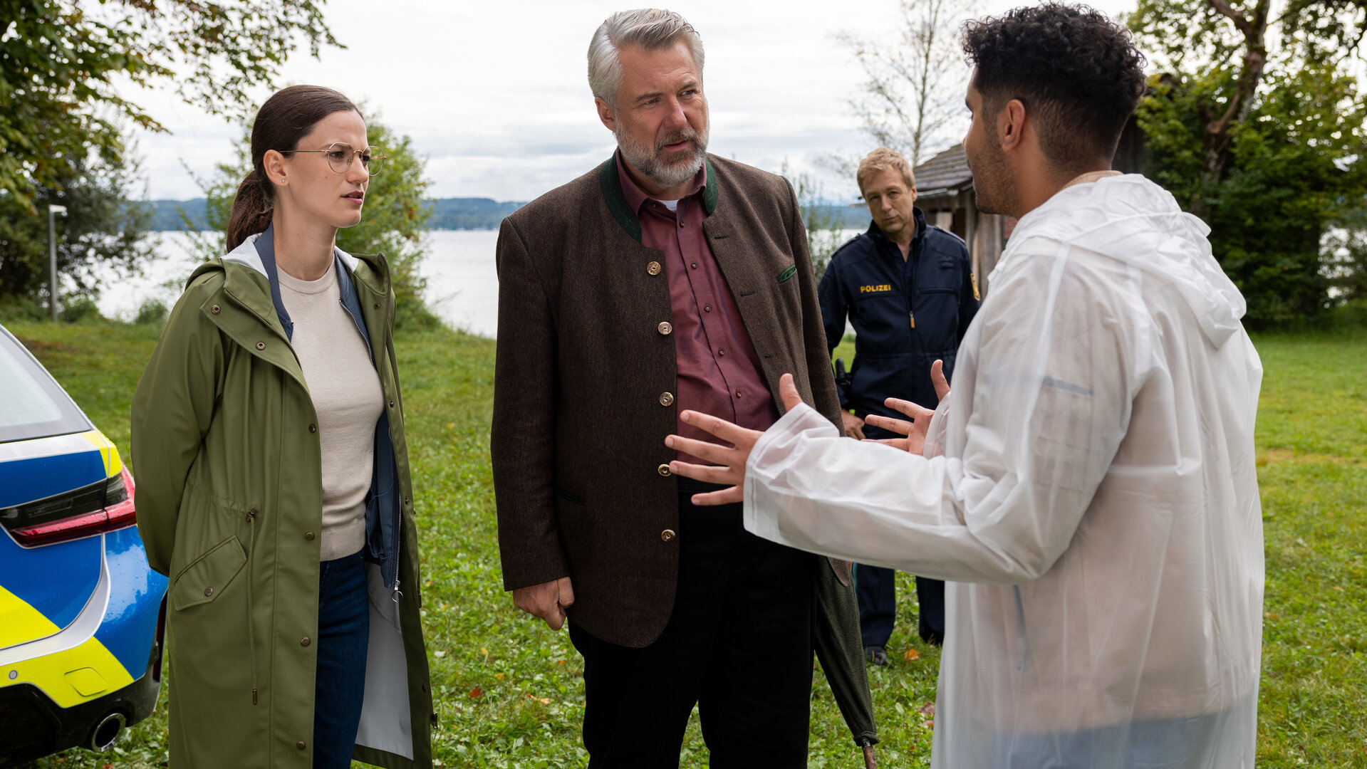 "Die Rosenheim-Cops - Der Steg ist weg": Die Hauptkommissare Beck (Michaela Weingartner) und Stadler (Dieter Fischer) stehen Dominik Meis (Younes Tissinte) gegenüber, der gestikulierend in einem weißen Regenmantel vor ihnen auf dem Rasen steht. Im Hintergrund verfolgt Polizeihauptmeister Mohr (Max Müller) das Geschehen. Neben ihnen ist das Heck eines Polizeifahrzeugs zu sehen, hinter ihnen ein See.