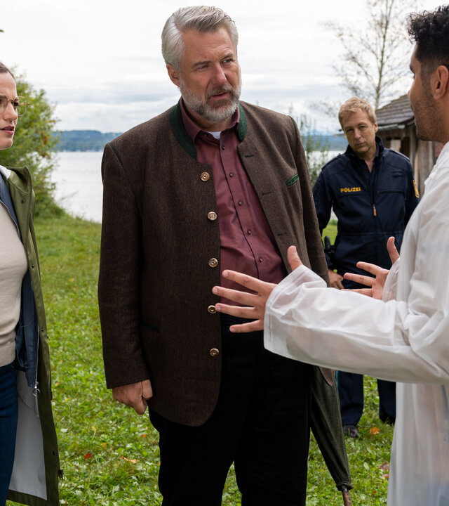"Die Rosenheim-Cops - Der Steg ist weg": Die Hauptkommissare Beck (Michaela Weingartner) und Stadler (Dieter Fischer) stehen Dominik Meis (Younes Tissinte) gegenüber, der gestikulierend in einem weißen Regenmantel vor ihnen auf dem Rasen steht. Im Hintergrund verfolgt Polizeihauptmeister Mohr (Max Müller) das Geschehen. Neben ihnen ist das Heck eines Polizeifahrzeugs zu sehen, hinter ihnen ein See.