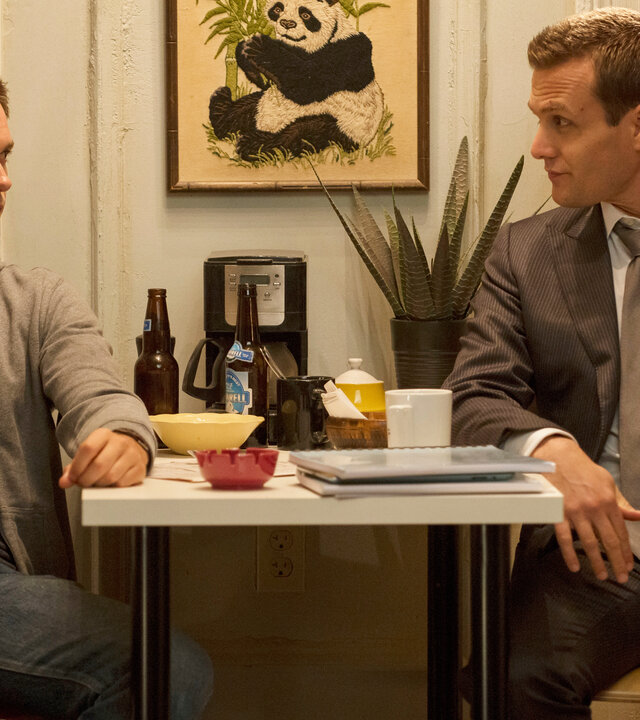 "Suits - Abrechnung": Mike (Patrick J. Adams) und Harvey (Gabriel Macht) sitzen sich in Mikes Küche gegenüber und blicken sich vertraut an. Auf dem Tisch zwischen ihnen stehen zwei Bierflaschen, Schalen und Unterlagen. Mike ist leger gekleidet, während Harvey einen Anzug trägt.