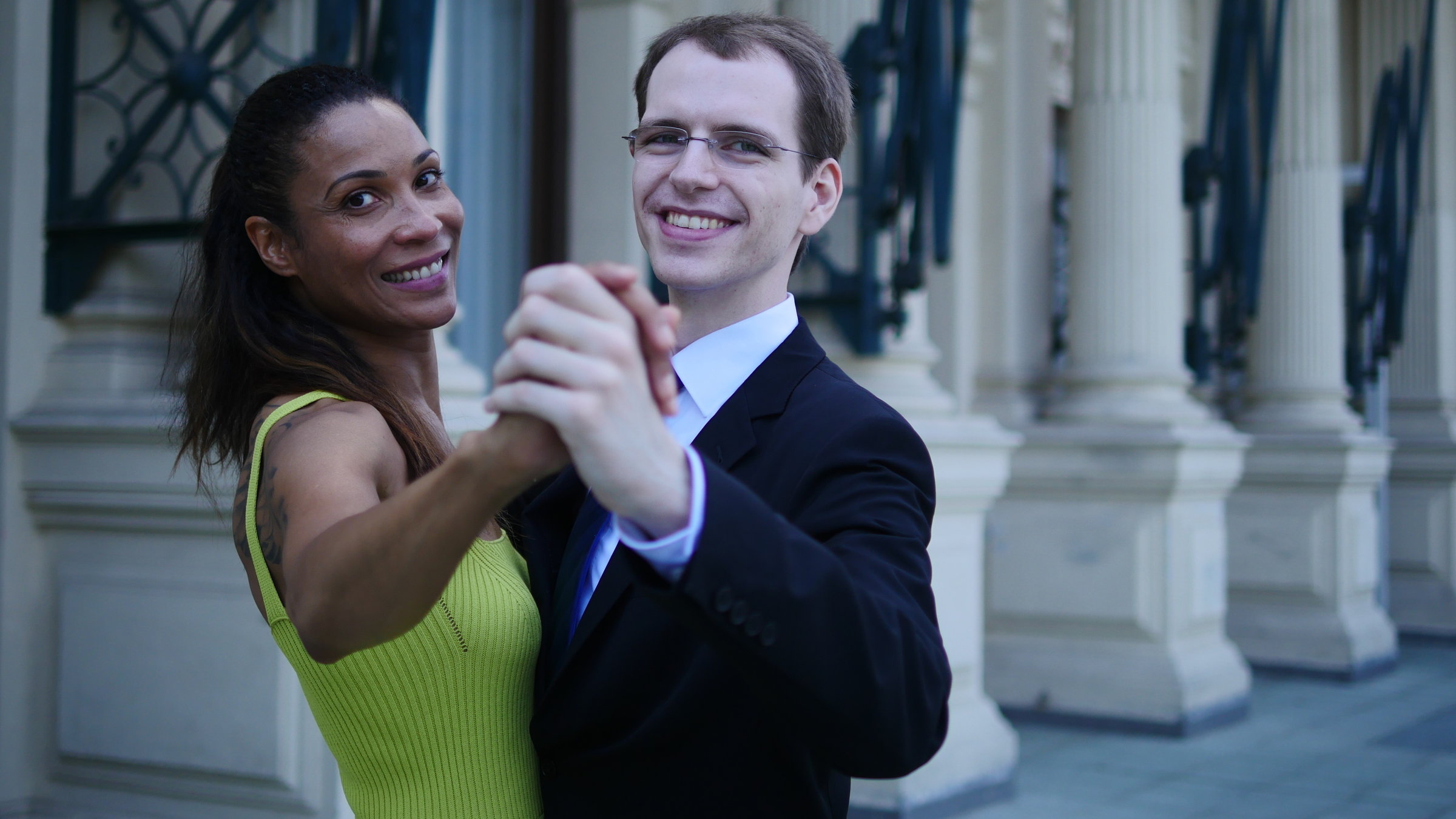 "Tanzwelten (2/2) - Wiener Walzer & Swing": Die Presenterin Annabelle Mandeng  im Halbprofil in einer Walzer Pose mit einem Tänzer. Im Hintergrund  sind weiße Säulen eines Gebäudes mit blauem Gitter