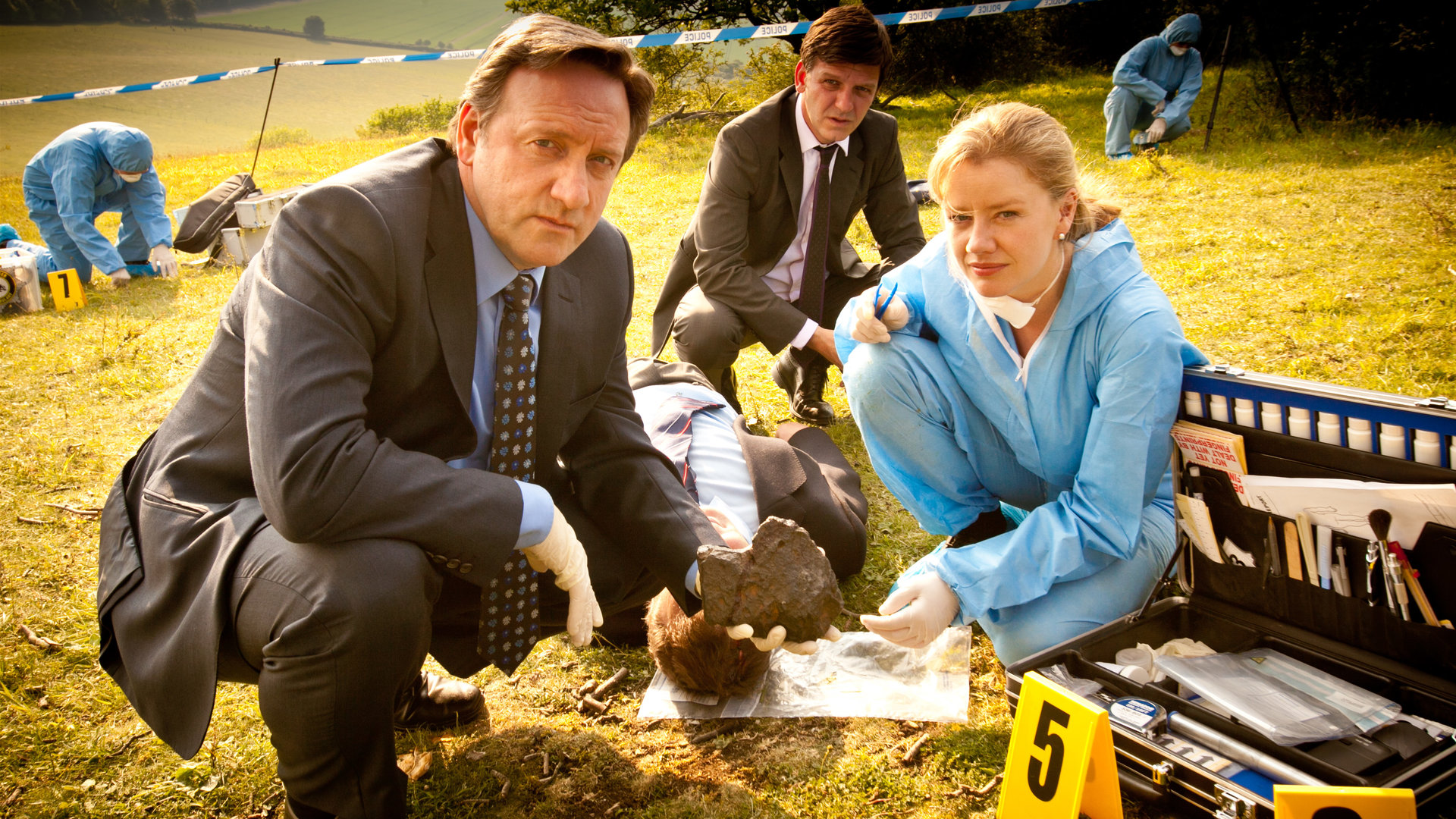 "Inspector Barnaby - Sonne, Mord und Sterne": Inspector Barnaby (Neil Dudgeon), Sergeant Ben Jones (Jason Hughes) und Kate Wilding (Tamzin Malleson) hocken um eine Leiche herum, die auf einer Wiese liegt. Im Hintergrund sind weitere Spurensicherer zu sehen.