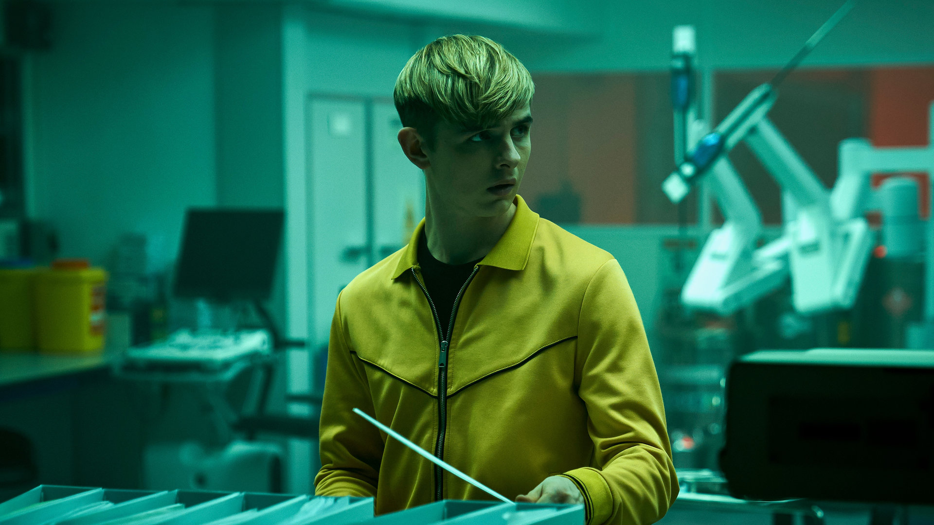 "Alex Rider - Verschwörung": Alex Rider (Otto Farrant) steht in einem grün beleuchteten Raum. Vor ihm steht eine Reihe von Ordnern. Alex blickt schockiert zur Seite.