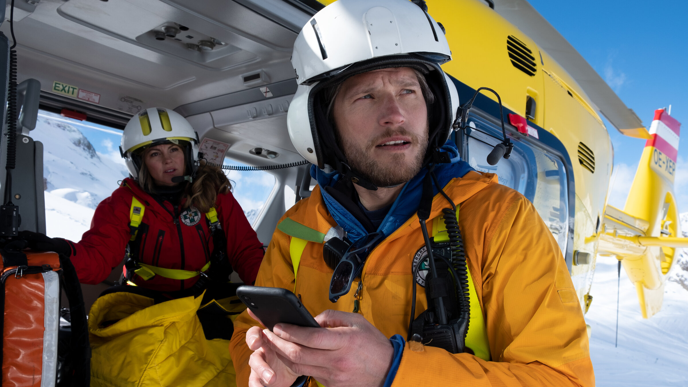 "Die Bergretter: Einfach hier bleiben":''In Schneelandschaft - Katharina (Luise Bähr) sitzt im Hubschrauber. Markus (Sebastian Ströbel) steht daneben und hält ein Mobiltelefon in der Hand. Beide tragen ihre Einsatzkleidung und schauen in die Ferne.