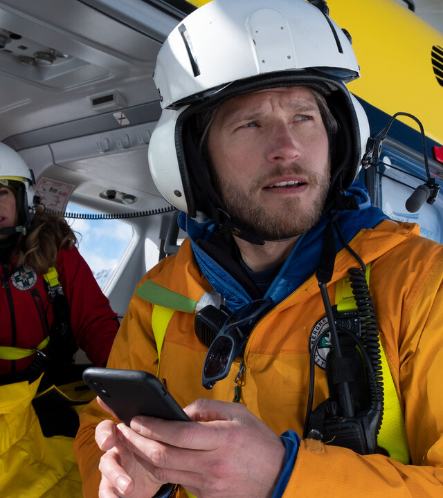 "Die Bergretter: Einfach hier bleiben":''In Schneelandschaft - Katharina (Luise Bähr) sitzt im Hubschrauber. Markus (Sebastian Ströbel) steht daneben und hält ein Mobiltelefon in der Hand. Beide tragen ihre Einsatzkleidung und schauen in die Ferne.