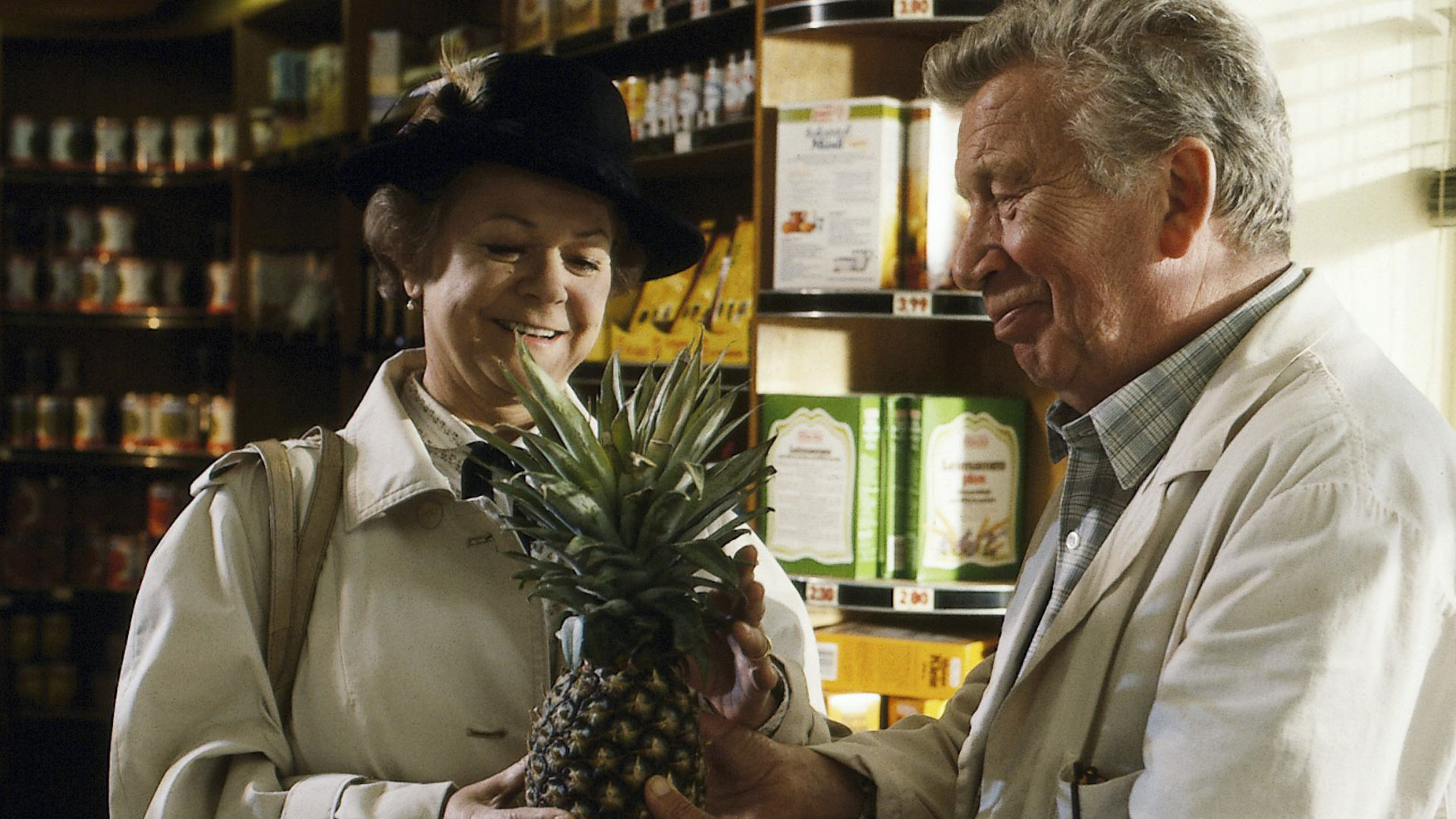 "Zwei Münchner in Hamburg - Familienzuwachs": Vadder Haack (Heinz Reincke) schenkt Fanny (Enzi Fuchs) eine Ananas. Sie stehen in einem kleinen Supermarkt, im Hintergrund sind befüllte Regale zu sehen.