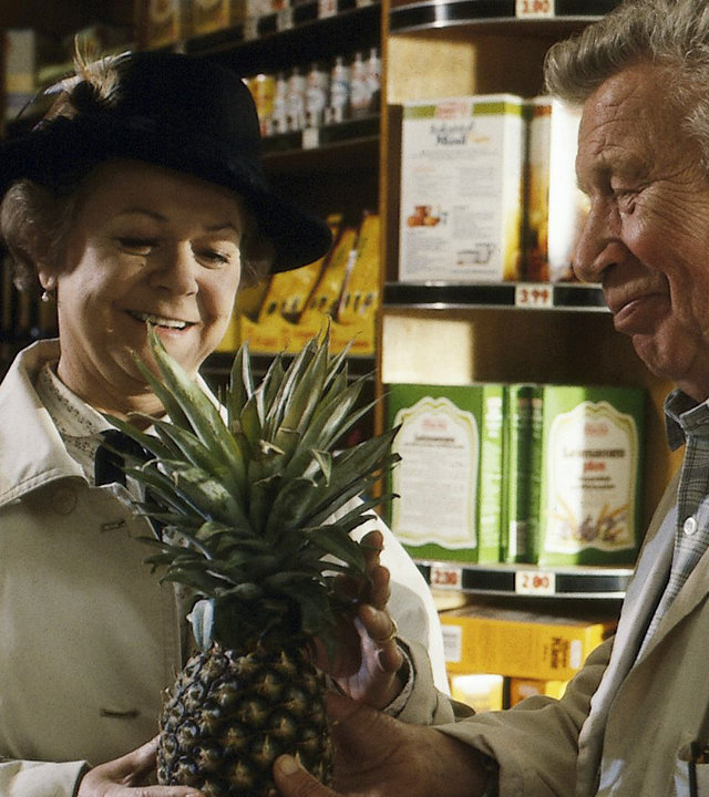 "Zwei Münchner in Hamburg - Familienzuwachs": Vadder Haack (Heinz Reincke) schenkt Fanny (Enzi Fuchs) eine Ananas. Sie stehen in einem kleinen Supermarkt, im Hintergrund sind befüllte Regale zu sehen.
