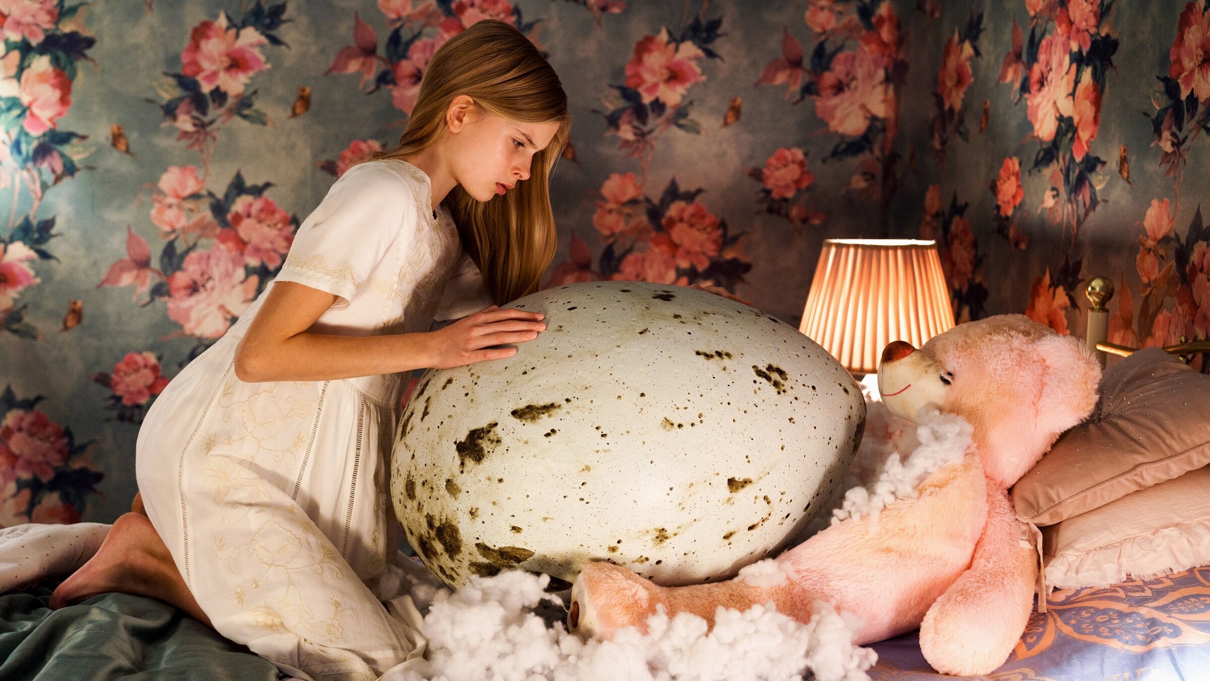 "Hatching": Halbtotale: Tinja (Siiri Solalinna) kniet auf Bett in weißem Nachthemd über einem überdimensionalen, gefleckten Ei, auf das sie mit neugierigem Blick ihre Hände gelegt hat. Die Wände des Kinderzimmers sind mit einer auffälligen Blumentapete tapeziert (rosa Blumen auf grauem Hintergrund).