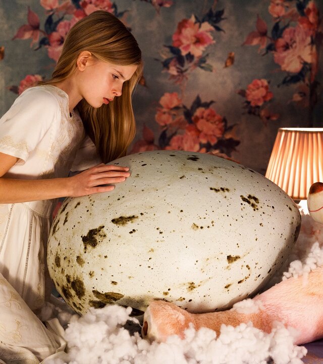 "Hatching": Halbtotale: Tinja (Siiri Solalinna) kniet auf Bett in weißem Nachthemd über einem überdimensionalen, gefleckten Ei, auf das sie mit neugierigem Blick ihre Hände gelegt hat. Die Wände des Kinderzimmers sind mit einer auffälligen Blumentapete tapeziert (rosa Blumen auf grauem Hintergrund).