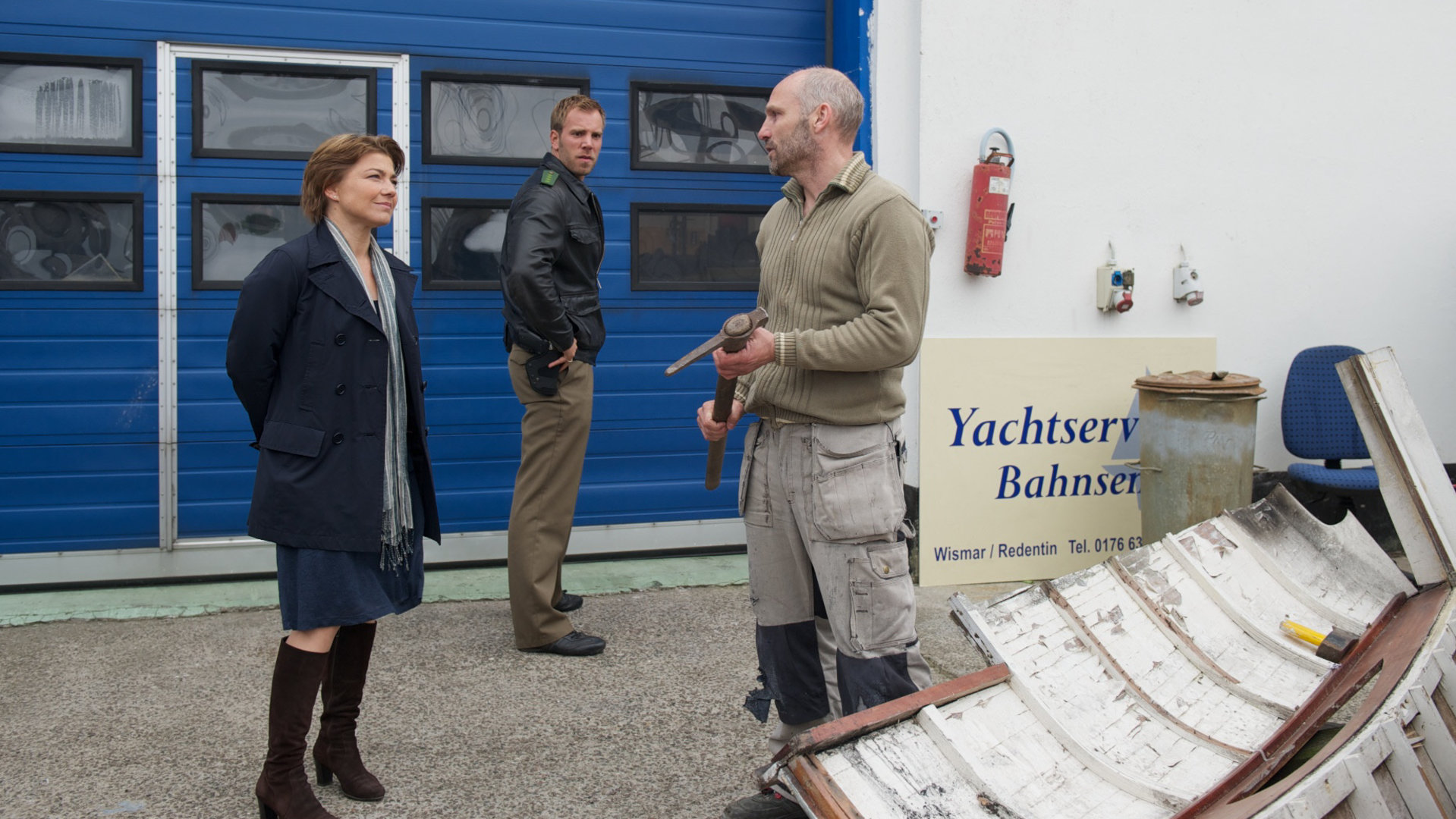 "SOKO Wismar - Väter und Söhne": Kommissarin Katrin Börensen (Claudia Schmutzler) und Kommissar Sven Herzog (Michale Härle) stehen mit Hajo Bahnsen (Thomas Wüpper) vor dem blauen Tor eines Yachtservices. Er hat einen Hammer in der Hand und ein verschrottetes Schiff neben sich liegen.