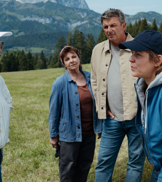 "Der Bergdoktor: Höhenflug": Auf einer Wiese vor einer Bergkulisse:  Mit schmerzverzerrtem Gesicht und leicht vorgebeugtem Oberkörper lehnt Charlie Eggers (Jaschka Lämmert) am Heck eines Segelflugzeugs. Ihr gegenüber steht Miriam Sterzinger (Carolina Vera), eine Sonnenbrille tragend, und redet mit Charly. Im Hintergrund stehen Dr. Martin Gruber (Hans Sigl) und Lisbeth Gruber (Monika Baumgartner) und beobachten die Frauen aufmerksam.